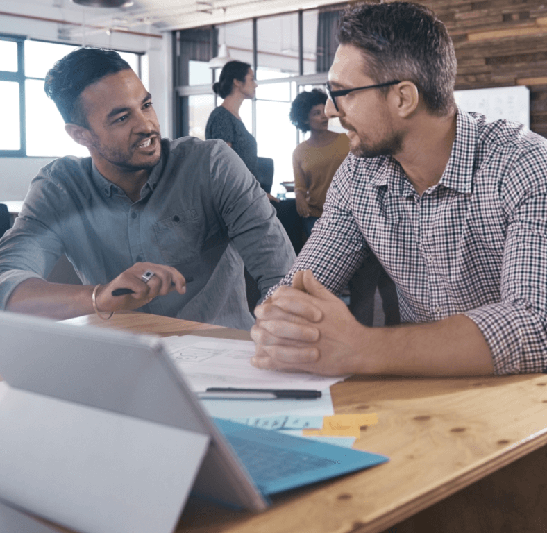 two males discussing project at table with documents and laptop